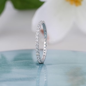 May include: A thin silver-toned metal band ring featuring a continuous row of small, clear, round-cut stones set along the outer circumference. The ring is positioned vertically, displaying a uniform, sparkling design with a polished metallic finish.