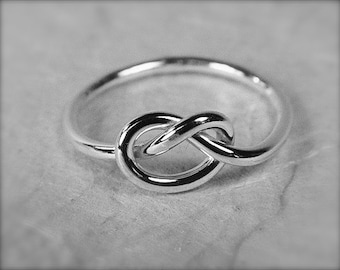 May include: A silver-colored ring featuring a knot design. The ring's band is smooth and circular, leading to the knot centerpiece. The knot is formed by the ring's metal, creating a continuous loop. The ring is shown in a close-up shot.