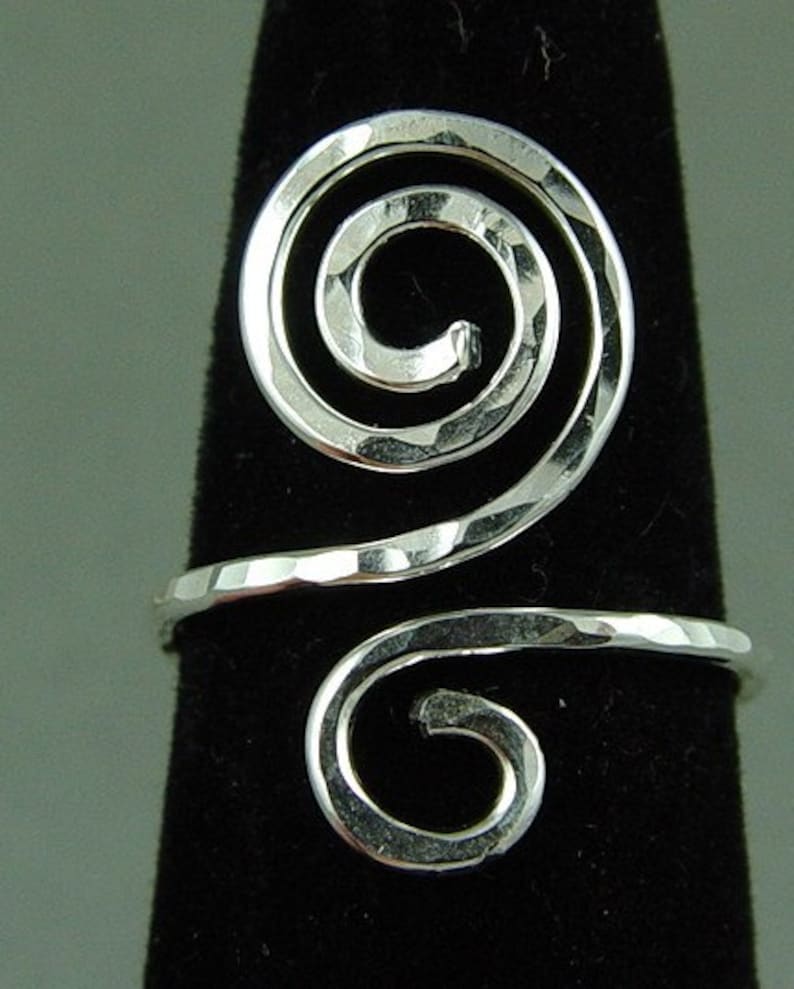May include: A silver-colored ring with a hammered texture. The ring features a spiral design, with the spiral wrapping around the finger. The ring is displayed on a black background.