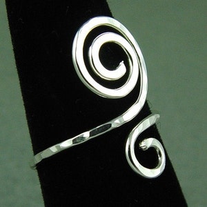 May include: A silver-toned, hammered-finish ring with a spiral design. The ring features a large, open spiral at the top and a smaller spiral at the bottom, connected by a band. The ring is displayed on a black surface.