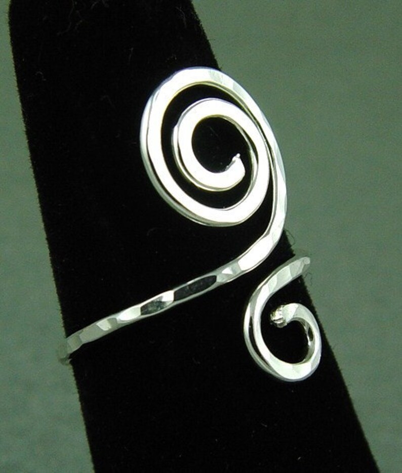 May include: A silver-toned, hammered-finish ring with a spiral design. The ring features a large, open spiral at the top and a smaller spiral at the bottom, connected by a band. The ring is displayed on a black surface.