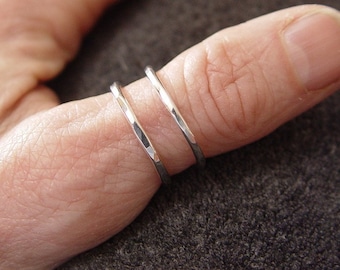 May include: Two silver rings with a hammered texture are worn on a finger. The rings are thin and have a slightly irregular surface, reflecting light. The rings are likely made of sterling silver and are a minimalist jewelry design.