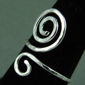 May include: A silver-toned ring with a hammered texture. The ring features a spiral design on the top and a curved band that wraps around the finger. The ring is displayed on a black surface.
