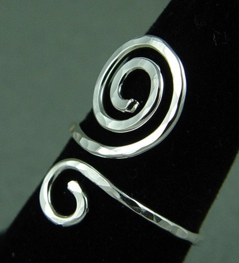 May include: A silver-toned ring with a hammered texture. The ring features a spiral design on the top and a curved band that wraps around the finger. The ring is displayed on a black surface.