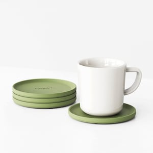 Può includere: Una tazza in ceramica bianca con manico poggia su un sottobicchiere rotondo verde oliva. Accanto, tre sottobicchieri impilati dello stesso colore. La parola "objekt" è stampata sul sottobicchiere superiore. Gli oggetti sono su una superficie bianca.