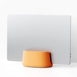 May include: A silver rectangular object is supported by an orange base. The base has a rounded shape. The background is white, creating a clean, minimalist aesthetic. The object appears to be a screen.