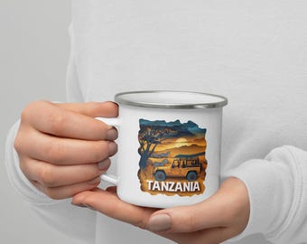 May include: White enamel mug with a safari scene featuring a vehicle, zebras, and a sunset. The word "TANZANIA" is printed in a bold, orange font at the bottom of the mug. The mug has a silver rim and a white handle.