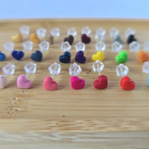 May include: A selection of heart-shaped stud earrings in a range of colours, including white, pink, red, blue, and green. The earrings are presented on a wooden tray, highlighting their diverse hues and designs. The earrings are small and delicate.