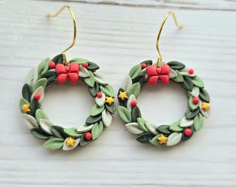 May include: A pair of dangle earrings featuring circular wreaths made of dark and light green leaves. Each wreath is decorated with a small red bow, tiny red berries, and yellow star-shaped accents, suspended from gold-toned metal hooks.