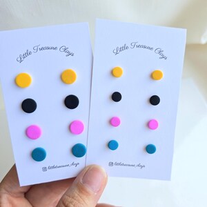 May include: Two white display cards labelled Little Treasure Clays, each holding four pairs of circular stud earrings in yellow, black, pink, and teal. The earrings are arranged in vertical columns on the cards, which also feature an Instagram icon.