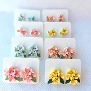 Puede incluir: Una colección de ocho pares de pendientes de botón presentados en tarjetas blancas. Cada par presenta pequeños racimos de flores en varios colores como azul claro, rosa y amarillo, con centros dorados y pequeñas hojas verdes en la base.