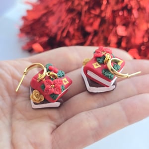 Op de afbeelding: Een paar feestelijke oorbellen met kerstthema. Elke oorbel heeft een miniatuur rood cake-ontwerp met witte glazuur, gouden accenten en details van poinsettia's en rozen. De oorbellen hebben goudkleurige oorringsluitingen.