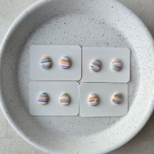 May include: Six pairs of colourful striped egg-shaped earrings on white cards. The earrings are pastel pink, yellow, blue, and purple.