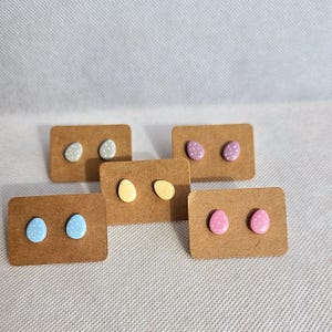 May include: Six pairs of small, egg-shaped earrings on brown card stock. The earrings are pastel colours with white polka dots. The colours are light blue, yellow, pink, and grey.