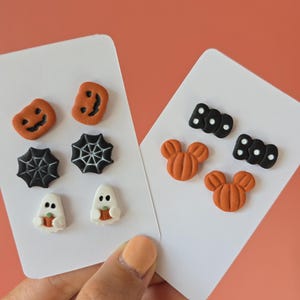 Può includere: Orecchini a bottone a tema Halloween su cartoncini bianchi. Gli orecchini presentano disegni di zucca, ragnatela, fantasma e Topolino in arancione, nero e bianco. La parola "BOO" è presente anche in borchie nere.