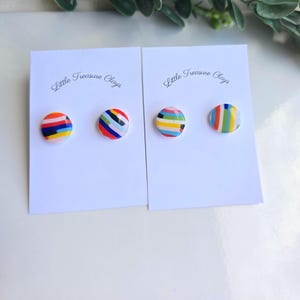 May include: Two white display cards labelled Little Treasure Clays, each holding a pair of round stud earrings. The earrings feature multicoloured horizontal and diagonal stripes in shades of red, blue, yellow, green, and black on a smooth surface.
