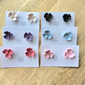 Op de afbeelding: Een verzameling bloemvormige oorstekers in verschillende kleuren, waaronder wit, zwart, paars, lichtblauw, rood en roze. Elk paar wordt gepresenteerd op een witte kaart met een kleine illustratie. De oorbellen hebben een goudkleurig midden.