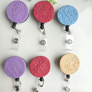 May include: Six circular badge reels arranged in two rows. The top row features purple, red, and blue reels with embossed patterns. The bottom row includes a purple, red, and beige reel, each with distinct textured designs and clear plastic straps.