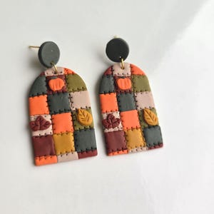 May include: A pair of arch-shaped dangle earrings featuring a patchwork quilt design in shades of orange, dark grey, brown, mustard yellow, and beige. Each earring has a small dark grey circular stud top and embossed pumpkin and leaf details.