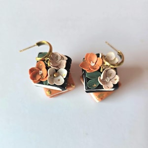 May include: A pair of floral earrings. Each earring features a black square with orange and white flowers, green leaves, and gold accents. The earrings have gold-coloured hoop closures.