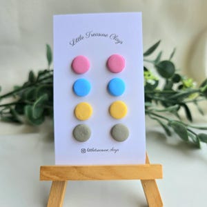 May include: A set of eight circular stud earrings arranged in four pairs on a white display card. The colours include pink, blue, yellow, and grey. The card features the text Little Treasure Clays and an Instagram icon with the handle littletreasure_clays.
