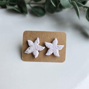 Puede incluir: Un par de pendientes de botón en forma de estrella blanca con una superficie texturizada y pequeñas motas doradas incrustadas. Los pendientes están sujetos a una tarjeta rectangular marrón, cada estrella presenta cinco puntas.