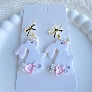 Könnte beinhalten: Weiße ohrringe in Hasenform mit rosa Akzenten und goldfarbenen Schleifen. Die Ohrringe haben ein Blumendesign an der Basis und sind an einer weißen Karte befestigt. Ein bezauberndes Accessoire für Ostern oder den Frühling.