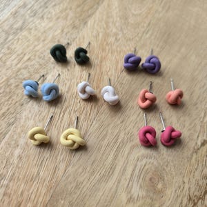 May include: A collection of colorful knot-shaped stud earrings. The earrings are in various colors, including green, blue, white, yellow, orange, and pink. Each pair has a silver post. The earrings are displayed on a light brown wooden surface.
