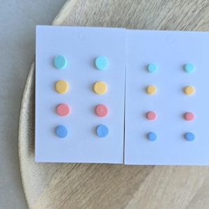 Op de afbeelding: Twee sets pastelkleurige oorstekers op witte kaarten. Elke kaart bevat zes ronde oorbellen in tinten blauw, geel, roze en groen. De oorbellen worden gepresenteerd op een houten dienblad.