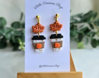 May include: A pair of dangle earrings on a white display card labeled Little Treasure Clays. Each earring features an orange maple leaf charm above a white coffee cup with a black lid, a brown sleeve, and a small orange pumpkin detail.