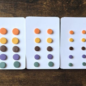 Puede incluir: Tres tarjetas blancas que muestran pendientes de botón coloridos. Cada tarjeta presenta seis pares de pendientes en naranja, amarillo, marrón, morado y verde. Los pendientes están dispuestos verticalmente, con tamaños decrecientes de izquierda a derecha.