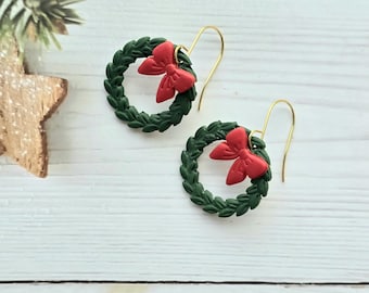 May include: A pair of dangle earrings featuring circular dark green wreaths with textured foliage details. Each wreath is accented with a small, bright red bow attached to the top. The earrings hang from thin, curved gold-toned metal hooks.