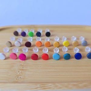 Peut inclure: Une collection de boucles d'oreilles à clous colorées dans diverses teintes, dont blanc, rose, rouge, orange, jaune, vert, bleu et noir. Les boucles d'oreilles sont disposées sur un plateau en bois, mettant en valeur leurs couleurs et leurs designs variés.
