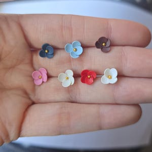 May include: A selection of small, flower-shaped stud earrings in shades of pink, white, red, blue, and brown. Each earring has a gold-coloured centre. The earrings are presented against a light background.