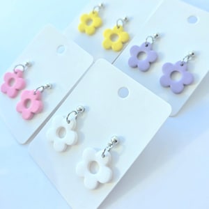 May include: A selection of flower-shaped earrings in pink, yellow, white, and lilac. Each earring is suspended from a silver-coloured hook and presented on a white card. The earrings feature a simple, minimalist design, suitable for everyday wear.