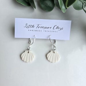 May include: A pair of white seashell-shaped charms hanging from silver-toned hoop earrings. The earrings are attached to a white rectangular display card featuring the black text Little Treasure Clays HANDMADE TREASURES printed on the front.