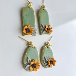 May include: Two pairs of dangle earrings featuring sage green arched and teardrop-shaped pendants. Each piece is decorated with a small yellow sunflower with a dark brown centre and grey leaves. The edges are trimmed in gold, and each earring has a gold post.