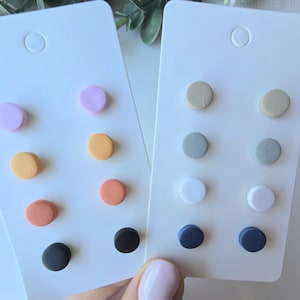 Peut inclure: Deux présentoirs à boucles d'oreilles blancs avec des ensembles de clous d'oreilles ronds et colorés. L'une des cartes présente des boucles d'oreilles dans les tons de violet, orange, corail et noir. L'autre carte a des boucles d'oreilles beiges, grises, blanches et bleu marine.