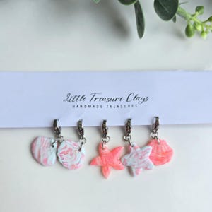 Könnte beinhalten: Fünf Anhänger an einer weißen Präsentationskarte mit der Aufschrift Little Treasure Clays Handmade Treasures. Die Anhänger umfassen zwei rosa-weiß marmorierte Muscheln, einen einfarbig rosa Seestern, einen rosa-weiß marmorierten Seestern und eine rosa Muschel.