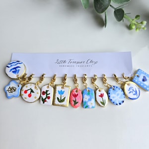 Pode incluir: Uma coleção de onze pequenos pingentes de argila presos a um cartão de exibição branco com a etiqueta Little Treasure Clays Handmade Treasures. Cada pingente apresenta designs pintados à mão, incluindo flores, cerejas e padrões em azul, vermelho e verde.