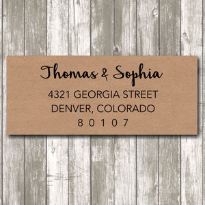 May include: A rectangular, kraft paper address label with the names "Thomas & Sophia" in elegant script. Below, the address reads "4321 GEORGIA STREET, DENVER, COLORADO 80107" in a simple, clean font. The label is set against a weathered, gray wooden background.