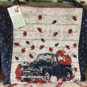 Pu&ograve; includere: Una borsa tote blu e bianca con un camioncino blu che trasporta zucche e foglie autunnali. La borsa ha un motivo a stelle blu e bianche sui lati e un bordo rosso e bianco.