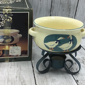 May include: A cream-colored ceramic bowl with two handles and a teal rim, featuring a graphic of two white geese with blue bows on a teal oval background. The bowl rests on a dark gray metal stand designed to hold a tea light candle.