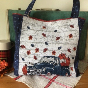 Pu&ograve; includere: Una borsa tote bianca con un camioncino blu che trasporta zucche e foglie autunnali. La borsa ha un motivo a stelle blu e bianche sui lati.