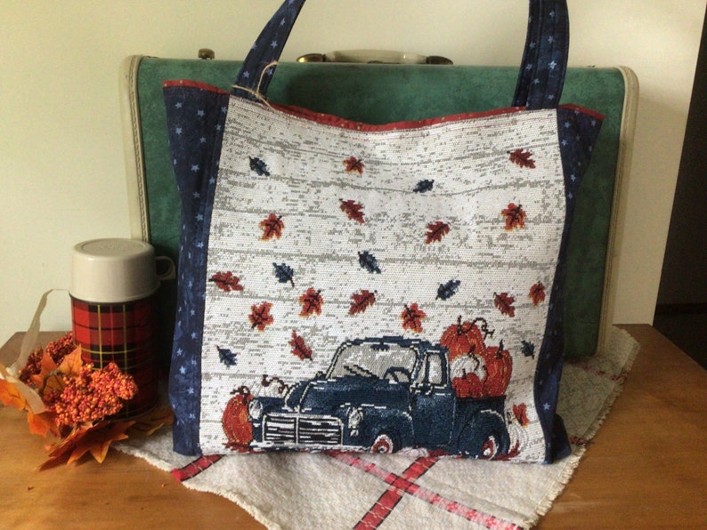 Pu&ograve; includere: Una borsa tote bianca con un camioncino blu che trasporta zucche e foglie autunnali. La borsa ha un motivo a stelle blu e bianche sui lati.