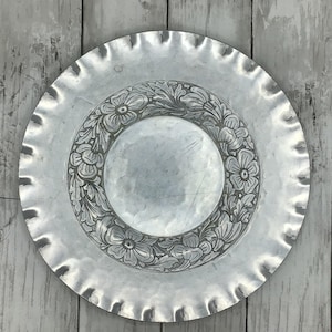 May include: A round silver-colored metal plate featuring a smooth center surrounded by a wide, embossed border decorated with a detailed floral and leaf pattern. The outer edge of the plate has a distinct, wavy, scalloped rim design.