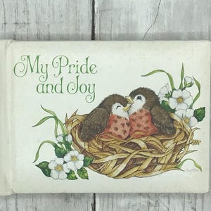 May include: A white card with the text "My Pride and Joy" and a painting of two birds in a nest with white flowers.