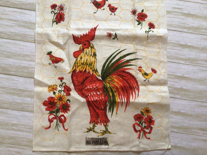 May include: A white tea towel with a red rooster and yellow chicks, red poppies, and a yellow honeycomb pattern. The text "Guaranteed All Pure Linen" is printed at the bottom.