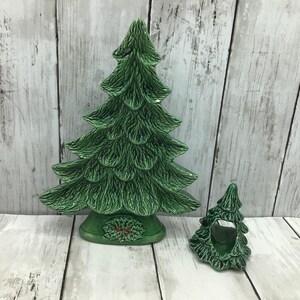 May include: Two green ceramic Christmas tree decorations. The larger tree has a detailed textured design and sits on a base with a red and green floral design. The smaller tree is a candle holder. Both are on a white wooden surface.