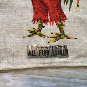 May include: A white linen tea towel with a rooster design and a silver label that reads "Parisian Prints All Pure Linen".